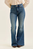 A person wearing medium-wash blue flare jeans with a light beige blouse and brown shoes, shown from the waist down against a plain light beige studio background.