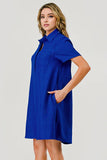 A woman modeling a short-sleeve collared shirt dress in a vibrant royal blue color with a subtle textured pattern, set against a plain light gray studio background.