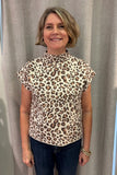 A woman with short blonde hair wearing a beige leopard-print mock neck top and dark blue jeans, standing in front of a gray curtain.