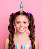 A young girl with brown pigtails and a rainbow unicorn horn headband, wearing a colorful striped top against a solid pink background.