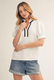 Woman wearing a white blouse with navy accents and denim shorts on a plain background
