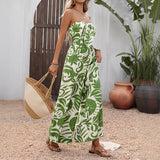 Woman wearing a strapless green leaf-patterned jumpsuit holding a straw bag outdoors.