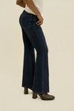 A person wearing dark blue flared jeans with a white top and brown block-heel boots, shown from the waist down against a plain beige studio background.