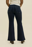 Rear view of a person wearing dark blue flare jeans with a cream top, standing against a plain beige studio background.