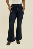 A person wearing dark blue flare jeans with a white top and brown boots, standing against a plain beige studio background.