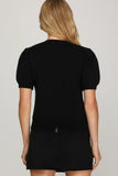 A woman viewed from the back wearing a black short-sleeve knit top or dress with a small silver zipper detail at the back hem, against a light gray studio background.