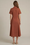 A woman viewed from the back wearing a short-sleeve collared rust-orange dress with a fitted waist and flared skirt, paired with beige heeled sandals, against a plain warm beige studio background.