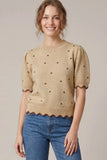 A woman wearing a beige short-sleeve knit sweater with brown polka dots and scalloped edges, paired with blue jeans, against a plain light beige studio background.