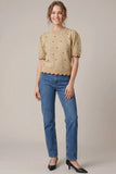 A woman modeling a beige short-sleeve knit top with small dark embroidered dots and a scalloped hem, paired with blue jeans and black heels, against a plain beige studio background.