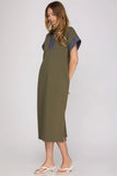 A woman modeling a short-sleeved olive green midi dress with contrasting dark blue fabric on the collar and sleeve cuffs, paired with white heeled thong sandals against a light gray studio background.