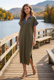 A woman stands on a wooden boardwalk by a lake with mountains and autumn trees in the background, wearing an olive green midi dress with a blue striped collar and short sleeves.