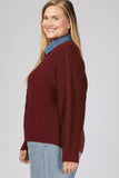 A woman wearing a burgundy cable-knit sweater layered over a blue denim collared shirt and light-wash jeans, shown from the side against a light gray studio background.