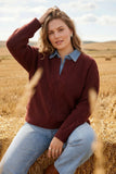 A woman sitting on a hay bale in a golden field, wearing a burgundy cable-knit sweater over a light blue collared shirt and light-wash jeans.