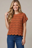 A woman with long blonde hair wearing a rust-orange short-sleeve knit top with a wavy textured pattern and blue jeans, standing against a solid beige studio background.