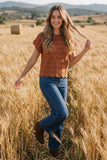 A woman stands in a golden wheat field wearing a rust-orange textured short-sleeve top, blue jeans, and brown boots.