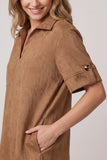 A close-up of a woman wearing a tan faux-suede short-sleeve collared dress or tunic with a partial placket and a decorative gold-toned button detail on the sleeve cuff, set against a plain light gray background.