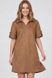 A woman wearing a brown faux-suede short-sleeve polo dress with rolled cuffs, photographed against a light gray studio background.