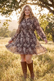 A woman stands in a sunlit grassy field wearing a long-sleeve mini dress with a brown and beige paisley pattern and brown knee-high boots.