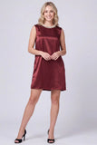 A woman modeling a sleeveless, knee-length satin dress in a deep burgundy color, styled with a pearl collar and black high heels against a light gray studio background.