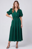 A woman modeling a dark green midi wrap dress with short puff sleeves, a V-neckline, and a tiered skirt, paired with black strappy heels against a light gray studio background.