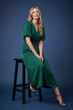 A woman wearing a green midi dress with puff sleeves sits on a black stool against a dark blue studio background.