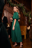 A woman wearing a dark green midi dress with puff sleeves and a tiered skirt stands in a decorated indoor setting with a wooden staircase, holiday garlands, and candles.