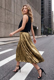 A woman walking on a city street crosswalk wearing a black sleeveless top and a shiny metallic gold pleated midi skirt, paired with black high-heeled sandals.