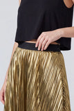 A close-up of a model wearing a shiny gold pleated skirt with a black waistband, paired with a black sleeveless top, against a light gray studio background.