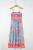 A sleeveless midi dress with thin straps, featuring a turquoise blue base with pink and red floral patterns and a smocked bodice, hanging on a wooden hanger against a white wall.