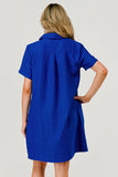 A woman shown from the back wearing a bright royal blue short-sleeve collared dress or tunic with a textured fabric, set against a plain light gray studio background.