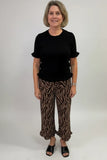 A woman modeling a black short-sleeve top with ruffled trim and brown-and-black zebra-print cropped pants, standing against a plain light gray studio background.
