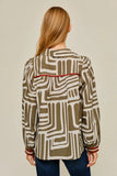 A woman viewed from the back wearing a long-sleeve button-up shirt with an abstract geometric pattern in olive green and cream, featuring thin red stripes at the cuffs and a red horizontal accent line across the upper back.