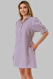 A woman modeling a short puff-sleeve shirt dress with vertical purple and white stripes, standing against a solid gray studio background.