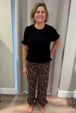 A woman standing indoors wearing a black short-sleeve top and brown pants with a black zebra-like pattern, posed in front of a light gray curtain.