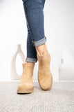 A person wearing tan suede ankle boots with a knit elastic side detail and cuffed blue jeans, standing on a light textured rug.