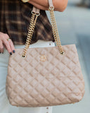 A close-up of a person holding a large quilted beige tote bag with gold chain shoulder straps and a gold monogram logo on the front.