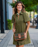 A woman wearing an olive green short-sleeve collared dress and carrying an olive green and brown crossbody bag with a chain strap, standing on a city sidewalk.