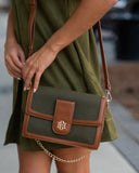A woman in an olive green dress carries a structured crossbody bag with an olive green body and brown leather trim, featuring a gold-tone monogram clasp and chain strap.