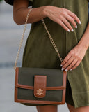 A woman wearing an olive green dress carries a structured shoulder bag with an olive green body, tan leather trim, and a gold chain strap. The bag features a front flap with a tan leather tab and a gold cross logo.