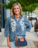 A woman wearing a blue denim shirt with white floral embroidery, light blue jeans, and a navy and brown crossbody bag, standing on a blurred city sidewalk.