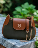 A brown and dark green leather shoulder bag with a gold-tone interlocking monogram logo on the front flap, resting on a gray stone surface with a blurred green garden background.
