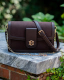 A dark brown leather crossbody bag with a gold-tone interlocking monogram logo on the front flap, resting on a gray stone ledge outdoors with green foliage in the background.