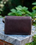 A dark brown suede shoulder bag with gold-tone chain strap details, resting on a gray stone ledge with a blurred green garden background.