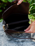 A dark brown leather shoulder bag with gold-tone chain detailing, shown open with a hand holding the flap and another hand pointing to the interior zippered compartment, set against a blurred outdoor background.