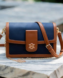 A navy blue and brown crossbody bag with a gold-tone monogram logo on the front flap, resting on a marble surface with a blurred outdoor background.