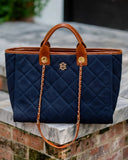 A navy blue quilted tote bag with brown leather handles and trim, featuring a gold-tone interlocking monogram logo and a chain strap, resting on a stone ledge.
