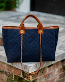 A navy blue quilted tote bag with tan leather handles and trim, featuring a gold-tone chain strap, resting on a stone ledge outdoors.
