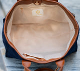 An open navy blue tote bag with tan leather trim, showing a cream-colored interior lining and a gold-tone 'CAROLINE HALL' logo patch on the inside pocket.
