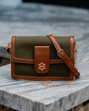A structured crossbody handbag with an olive green canvas body and brown leather trim, featuring a gold-tone monogram logo on the front flap and a gold chain strap, resting on a gray stone surface.