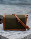 A rectangular shoulder bag with an olive green canvas body and brown leather trim, featuring a matching brown adjustable shoulder strap with a gold buckle and a small tassel detail, resting on a gray stone surface.
